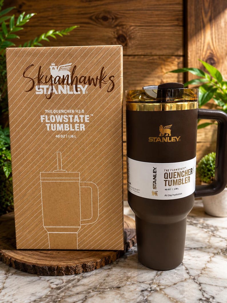 May include: A brown Stanley Flowstate Quencher Tumbler with a gold-colored rim and handle, next to a matching cardboard box. The box features the text "Skyanhawks" and "Stanley", along with the tumbler's details: "40 oz | 1.18 L".