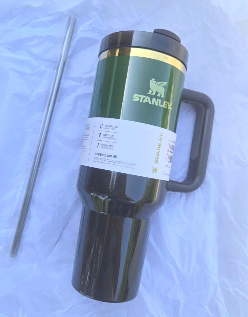 K&ouml;nnte beinhalten: Gr&uuml;n-schwarzer Stanley-Reisebecher mit Griff und silbernem Strohhalm. Der Becher hat einen goldenen Rand und das Stanley-Logo. Der Becher h&auml;lt Getr&auml;nke 11 Stunden kalt und 7 Stunden hei&szlig;.