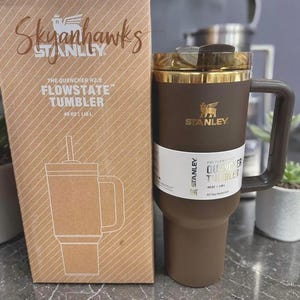 May include: A brown Stanley Flowstate tumbler with a gold-colored lid and handle, next to its matching box. The box features the text "Skyhawks" and a drawing of the tumbler. The tumbler holds 40 oz (1.18 L).