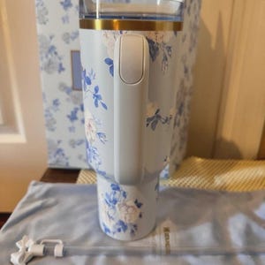 May include: A light blue tumbler with a floral design in white and blue. The tumbler has a handle, a gold rim, and a clear lid. A matching box and straw are also visible.