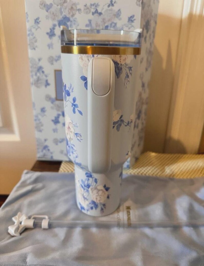 May include: A light blue tumbler with a floral design in white and blue. The tumbler has a handle, a gold rim, and a clear lid. A matching box and straw are also visible.
