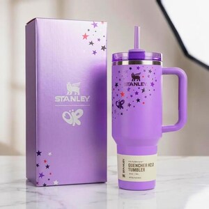 以下が含まれることがあります： 紫色のStanley Quencher H2.0タンブラー。ハンドルとストロー付き。お揃いの箱の隣にあります。タンブラーは星と蝶のデザインです。箱とタンブラーにはStanleyのロゴがあります。終日水分補給用に設計されています。