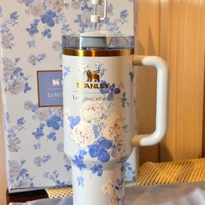 May include: A light blue Stanley tumbler with a floral design featuring white and blue roses. The tumbler has a white handle, a clear lid, and a bow on top. The box behind the tumbler has a matching floral pattern.