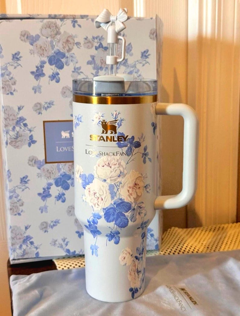May include: A light blue Stanley tumbler with a floral design featuring white and blue roses. The tumbler has a white handle, a clear lid, and a bow on top. The box behind the tumbler has a matching floral pattern.