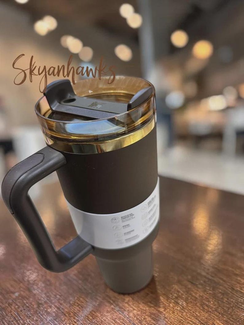 May include: A black and gold insulated travel mug with a handle. The mug has a clear lid with a black slider and a gold band around the top. The mug has a white band with symbols and text. The word "Skyanhawks" is written in cursive.