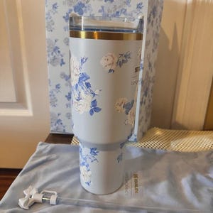 May include: A light blue Stanley tumbler with a floral design in white and blue. The tumbler has a gold rim and a clear lid. A matching box and a blue carrying bag with a straw are also present.
