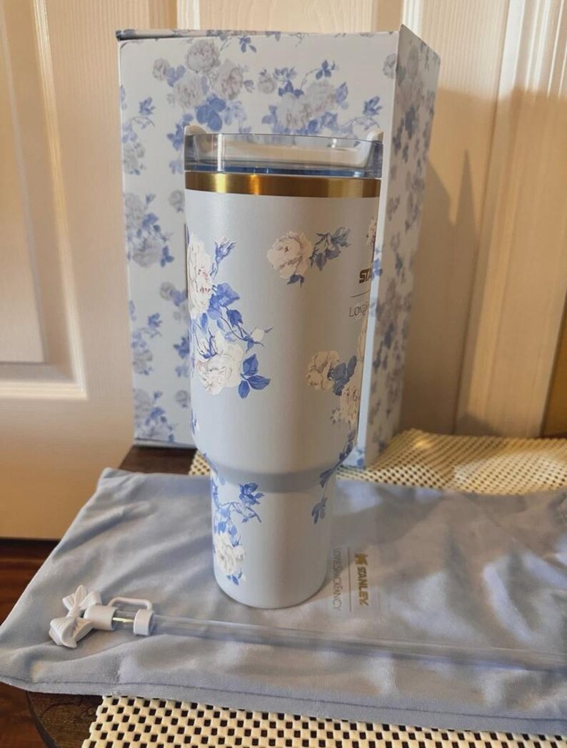 May include: A light blue Stanley tumbler with a floral design in white and blue. The tumbler has a gold rim and a clear lid. A matching box and a blue carrying bag with a straw are also present.