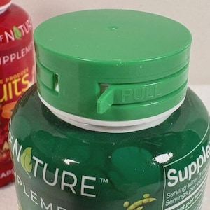 May include: Close-up of a green supplement bottle with a green cap. The cap has a "PULL" tab. The bottle is labelled "Nature Supplement" and contains a blend of fruits and vegetables.