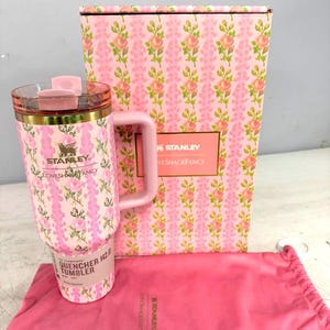 Op de afbeelding: Een roze Stanley Quencher H2.0 tumbler met een bloemenpatroon, een bijpassende roze handgreep en een doorzichtig deksel. De tumbler staat naast een roze doos met een bloemenpatroon en een roze trekkoordtas. De tumbler heeft de tekst "Stanley Loves Shack Fancy".