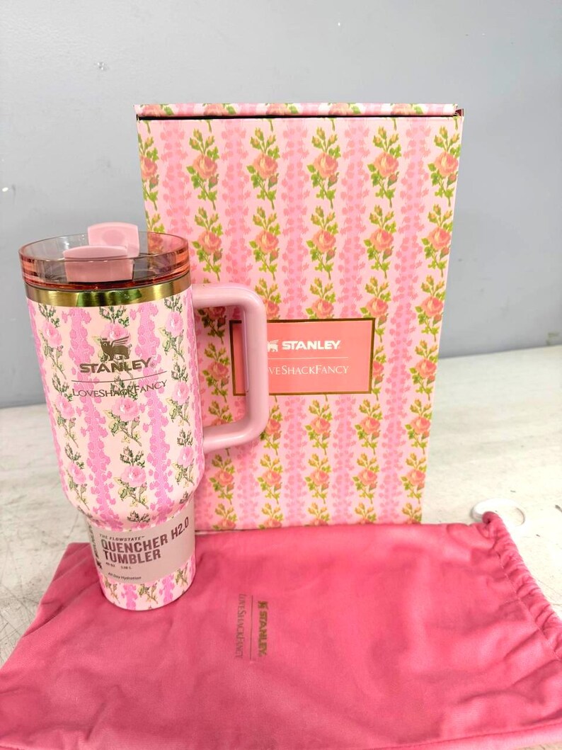 Op de afbeelding: Een roze Stanley Quencher H2.0 tumbler met een bloemenpatroon, een bijpassende roze handgreep en een doorzichtig deksel. De tumbler staat naast een roze doos met een bloemenpatroon en een roze trekkoordtas. De tumbler heeft de tekst "Stanley Loves Shack Fancy".
