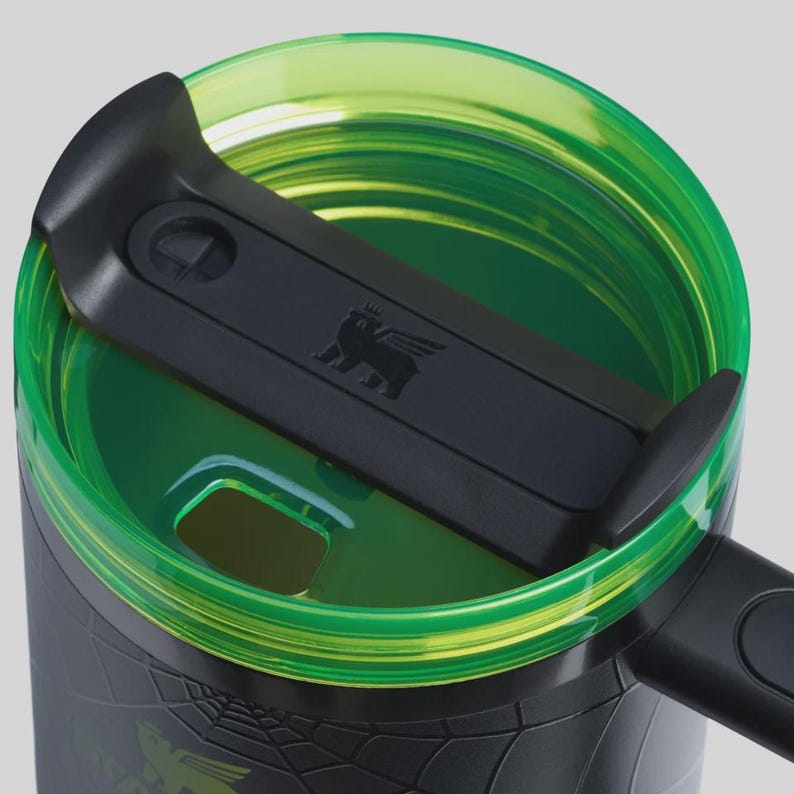 May include: Close-up of a black and green travel mug. The lid is black with a bear logo and a green rim. The mug's body is black with a textured pattern. The lid is open, revealing the interior.