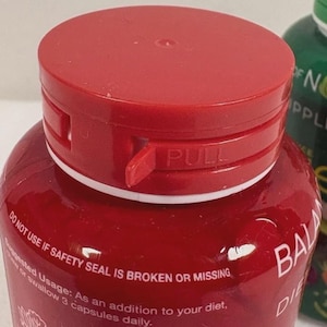 May include: A close-up of a red supplement bottle with a matching cap. The cap has a "PULL" tab. The bottle's label includes text about safety seals and suggested usage. The bottle is labeled "BALA DIE".