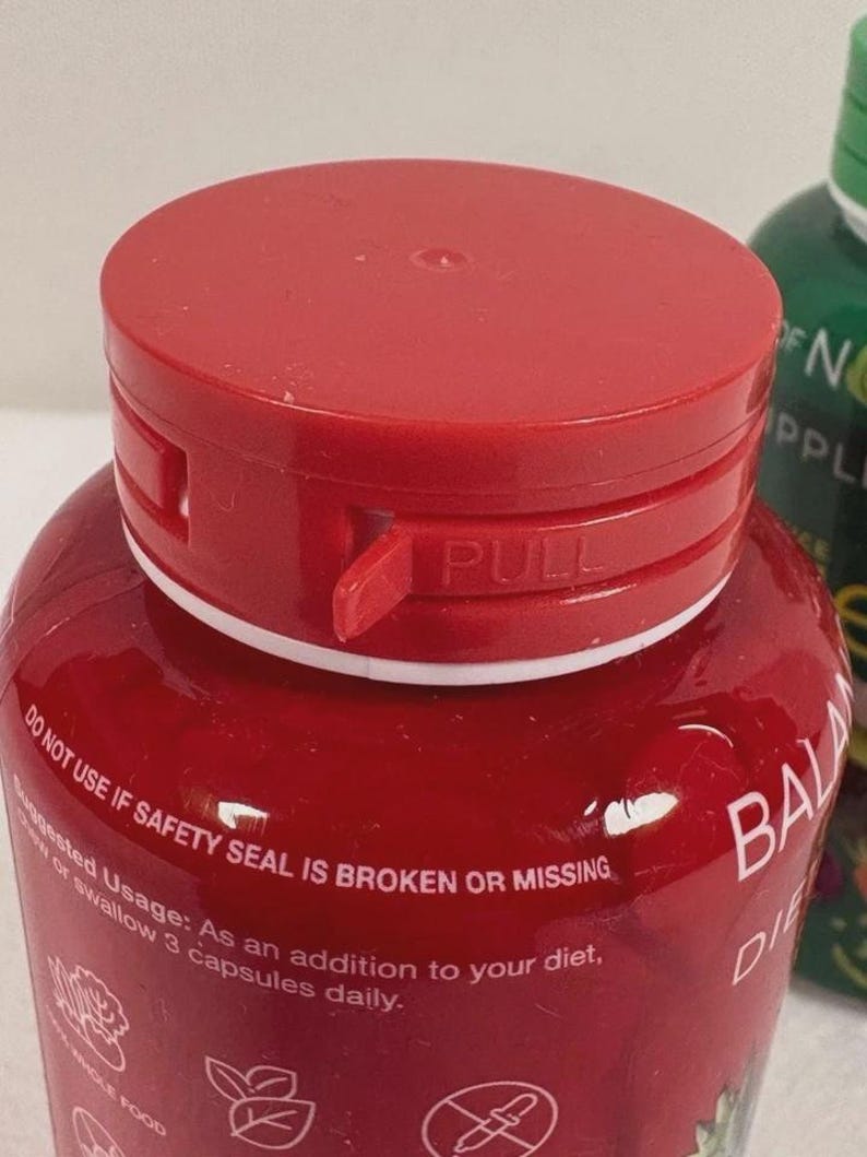 May include: A close-up of a red supplement bottle with a matching cap. The cap has a "PULL" tab. The bottle's label includes text about safety seals and suggested usage. The bottle is labeled "BALA DIE".