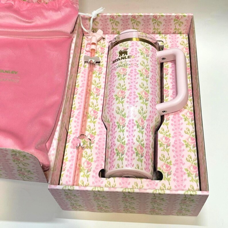Op de afbeelding: Een roze Stanley-beker met bloemenpatroon, handvat, rietje en deksel, gepresenteerd in een bijpassende geschenkverpakking. De beker heeft een herhalend patroon van roze en groene bloemen. De set bevat een roze trekkoordtas.