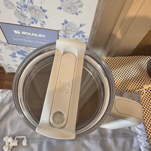 May include: A clear Stanley tumbler with a white lid and handle. The lid features the Stanley logo and a sliding closure. The tumbler is partially visible, with a light blue floral patterned box in the background. The text "STANLEY LOVESHACKFANCY" is visible on the box.