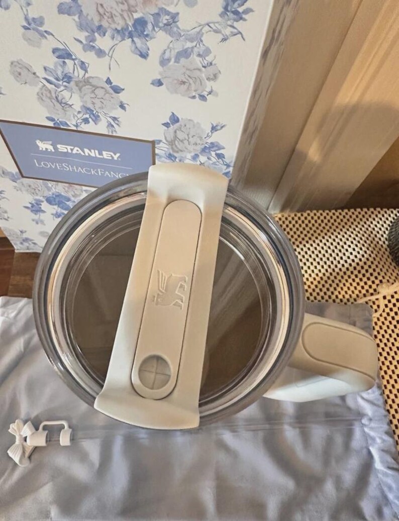 May include: A clear Stanley tumbler with a white lid and handle. The lid features the Stanley logo and a sliding closure. The tumbler is partially visible, with a light blue floral patterned box in the background. The text "STANLEY LOVESHACKFANCY" is visible on the box.