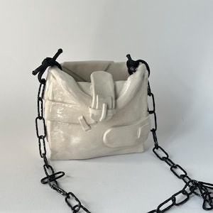 Peut inclure: Une sculpture de sac en céramique blanche présentant un design à rabat plié avec une sangle à boucle centrale. La pièce comprend une attache en cordon noir aux coins supérieurs et une longue chaîne en céramique noire texturée sur les côtés.