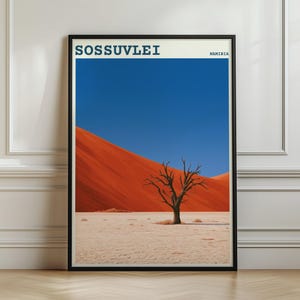 Op de afbeelding: Een ingelijste kunstdruk met een landschap van Sossusvlei, Namibië. De afbeelding toont een kale boom op een lichte woestijnbodem tegen een achtergrond van feloranje zandduinen en een heldere, diepblauwe lucht.