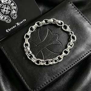 Op de afbeelding: Een zilveren armband met schakelketting ligt op een zwarte leren portemonnee. De armband heeft een karabijnsluiting. Een zwarte kaart met de tekst "Chrome Hearts" staat op de achtergrond.
