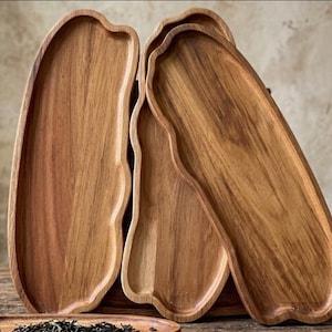 Könnte beinhalten: Eine Sammlung von Serviertabletts aus Holz mit organischen, fließenden Formen. Die Tabletts sind aus hellbraunem Holz gefertigt, das die natürliche Maserung zeigt. Das Design zeichnet sich durch einen erhöhten Rand und ein glattes, geschwungenes Innendesign aus.