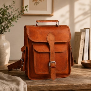 May include: A tan leather backpack featuring a top handle, a large front pocket with a flap, and a central vertical strap secured by a silver-toned metal buckle. The bag has side pockets and adjustable straps, showcasing a smooth, uniform texture.