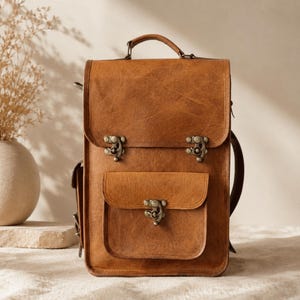 May include: A tan leather backpack featuring a rectangular silhouette, a top handle, and a fold-over flap secured by two antique-style metal latches. The front includes a smaller pocket with a matching metal latch closure and side pockets.