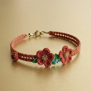 Pode incluir: Pulseira artesanal com uma faixa tecida castanho-avermelhada e um fecho dourado. Apresenta três aplicações de flores vermelhas com centros dourados e pequenos detalhes verdes e azuis. Comprimento: aprox. 18 cm.