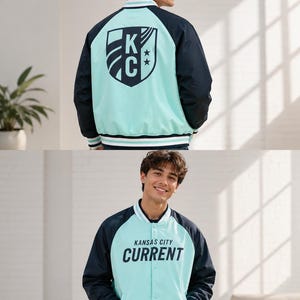 May include: A light blue and navy blue varsity jacket with the text "KANSAS CITY CURRENT" printed on the front. The back of the jacket features a shield design with the letters "KC" and two stars. The jacket has white and navy blue striped trim.