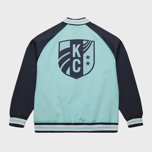 May include: A teal and navy blue bomber jacket with a shield logo on the back. The logo features the letters "KC" and two stars. The jacket has navy blue sleeves and white and teal striped trim on the collar, cuffs, and hem.