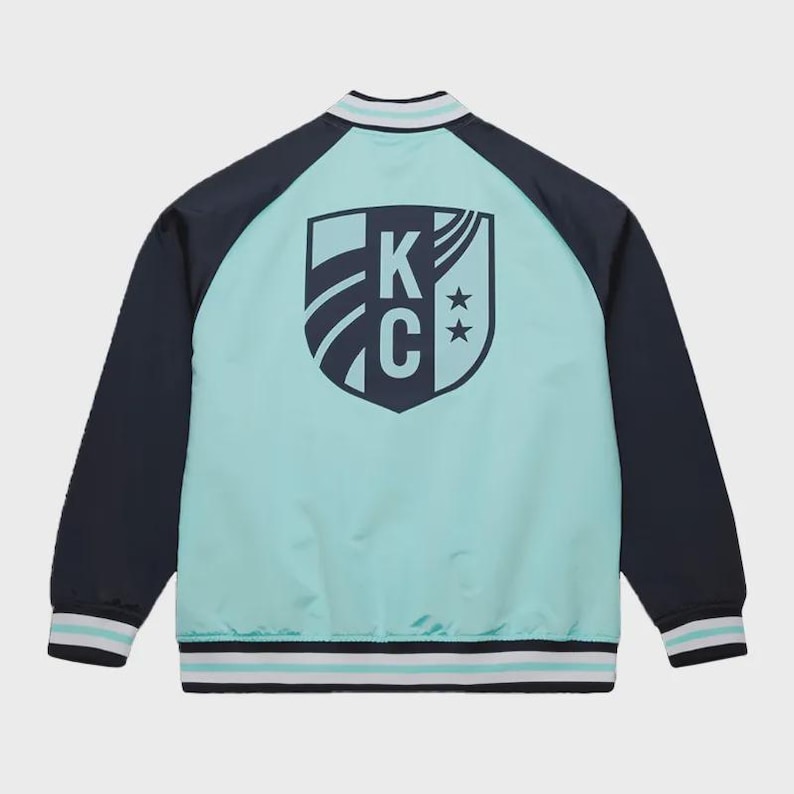 May include: A teal and navy blue bomber jacket with a shield logo on the back. The logo features the letters "KC" and two stars. The jacket has navy blue sleeves and white and teal striped trim on the collar, cuffs, and hem.