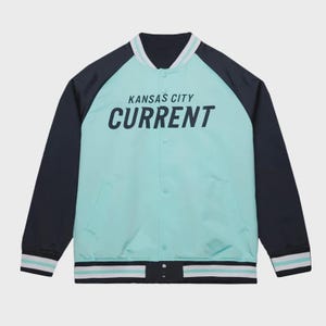 May include: A teal and navy blue varsity jacket with the text "KANSAS CITY CURRENT" in white. The jacket has a button-up front, side pockets, and striped detailing on the collar, cuffs, and hem.