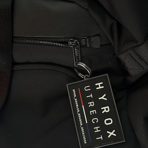 Puede incluir: Primer plano de una bolsa negra con cremallera y etiqueta. La etiqueta dice "HYROX UTRECHT" con el texto "OPEN DOUBLES SEASON 2025/2026". El tirador de la cremallera tiene un pequeño anillo de metal.