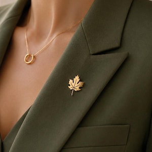 May include: A gold maple leaf brooch pinned to an olive green blazer. A delicate gold necklace with a circular pendant is also visible. The blazer has a classic cut and is worn over a matching top, creating a coordinated look.