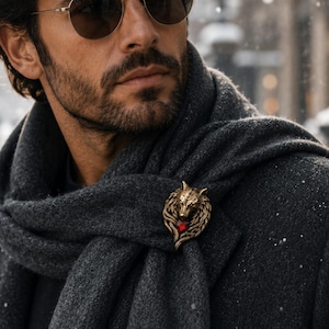 Puede incluir: Un broche de metal en tono dorado con forma de cabeza de lobo y una pequeña piedra preciosa roja brillante en la base, sujeto a una bufanda de lana gris carbón oscuro sobre un abrigo a juego durante una ligera nevada.