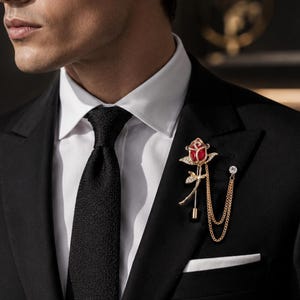 May include: A black suit with a white dress shirt and black tie. A gold-toned lapel pin in the shape of a red rose with a gold chain is attached to the suit's lapel. The pin has a small diamond accent.