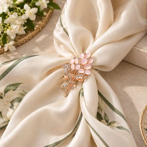 May include: A gold-toned metal scarf ring featuring a bouquet design with oval-shaped pale pink cabochon stones and small clear crystals. The brooch-style accessory is fastened to a cream-coloured silk scarf with a green leaf and floral pattern.