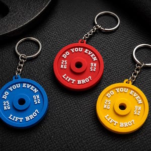 Peut inclure: Trois porte-clés circulaires en forme de disques de musculation de couleur bleue, rouge et jaune. Chacun présente le texte blanc en relief DO YOU EVEN LIFT BRO? et 25 KG. Les porte-clés sont fixés à des anneaux métalliques argentés par des chaînettes.