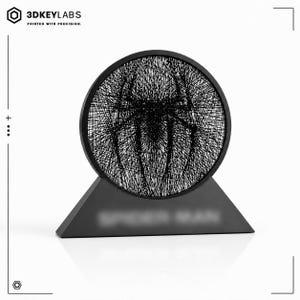May include: A black circular display piece featuring a complex web-like pattern forming a spider emblem in the center. The circular frame is mounted on a solid black triangular base with the text SPIDER-MAN printed on the front surface.