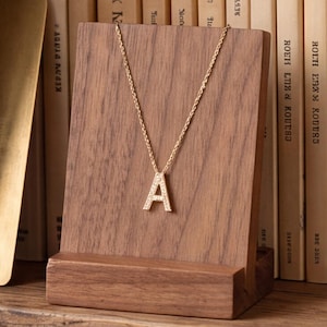 Puede incluir: Un collar de tono dorado con un colgante en forma de letra A incrustado con pequeñas piedras claras, exhibido en un soporte de madera marrón oscuro. El colgante cuelga de una cadena fina frente a una fila de libros en un estante.