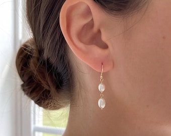 May include: A gold drop earring featuring two small, white, oval-shaped pearls suspended vertically from a thin gold hook. The pearls are smooth and lustrous, connected by a delicate gold wire, creating a simple and elegant dangling design.