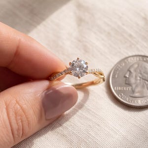 May include: A gold ring featuring a central round brilliant-cut stone held by six prongs, with a twisted band accented by small pavé-set stones. The ring is displayed next to a United States quarter coin for scale, showing its relative size.