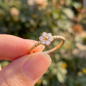 May include: A gold ring featuring a twisted rope-style band and a central flower-shaped cluster of six small, clear, round-cut stones surrounding a single gold bead at the center of the floral arrangement.