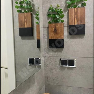 Può includere: Tre fioriere rettangolari in legno con un design bicolore, con una parte superiore in legno naturale e una parte inferiore nera. Ogni fioriera contiene piante verdi a foglia. Le fioriere sono montate su una parete piastrellata grigia, creando un'estetica moderna.