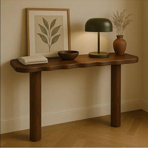 May include: A dark wood console table with a wavy edge, supported by two cylindrical legs. The table displays a framed botanical print, a small bowl, a folded cream-coloured cloth, a green lamp, and a brown vase with dried plants.