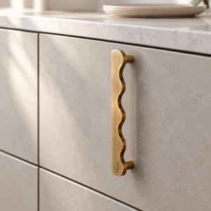 May include: A vertical brass cabinet handle with a wavy, scalloped edge on one side and a straight edge on the other. The handle features a brushed metallic finish and the word Albert is engraved near the bottom mounting post.