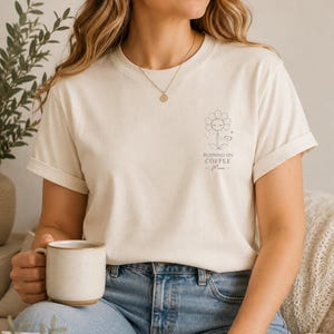 Könnte beinhalten: Ein cremefarbenes Kurzarm-T-Shirt mit einer kleinen schwarzen Strichgrafik auf der linken Brust. Das Design zeigt eine lächelnde Blume, die eine Kaffeetasse hält, mit dem Text "RUNNING ON COFFEE - Mom -" unterhalb der Blume gedruckt.