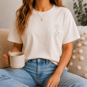 Könnte beinhalten: Ein cremefarbenes Kurzarm-T-Shirt mit einer kleinen schwarzen Strichzeichnung auf der linken Brust. Das Motiv zeigt eine schlafende Blume, die ein Kissen hält, mit dem Text "NO SLEEP CLUB" und "Mum" in einer einfachen Schrift darunter.