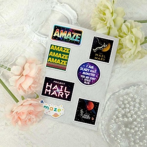 May include: A sticker sheet featuring space-themed designs including rectangular stickers with the text AMAZE and PROJECT HAIL MARY, a circular sticker with the text I AM SCARY SPACE MONSTER YOU ARE LEAKY SPACE BLOB, and a small cloud-shaped maze sticker.