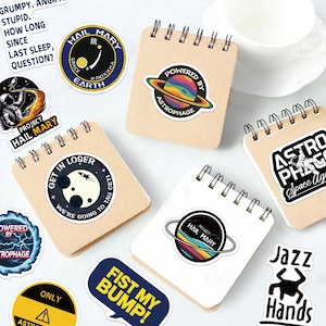 May include: A collection of square spiral-bound notebooks with kraft paper covers, decorated with various space-themed stickers including text like "Powered by Astrophage," "Get in loser, we're going to Tau Ceti," and "Project Hail Mary."