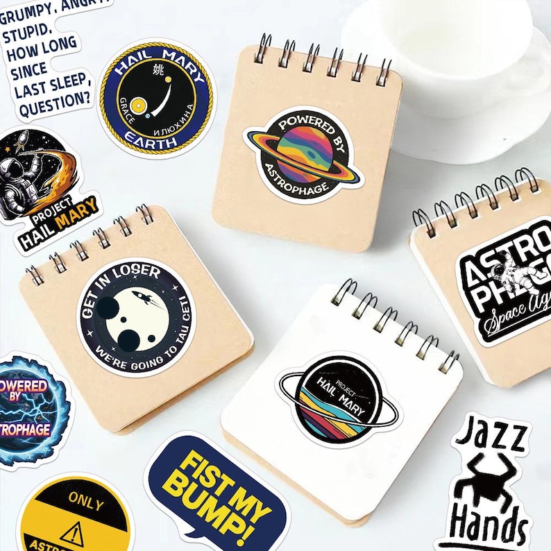 May include: A collection of square spiral-bound notebooks with kraft paper covers, decorated with various space-themed stickers including text like "Powered by Astrophage," "Get in loser, we're going to Tau Ceti," and "Project Hail Mary."